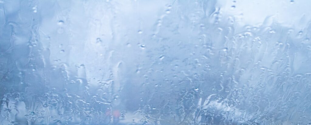 Close-up photograph of water droplets forming condensation on a home window, symbolizing the high indoor humidity that ove...