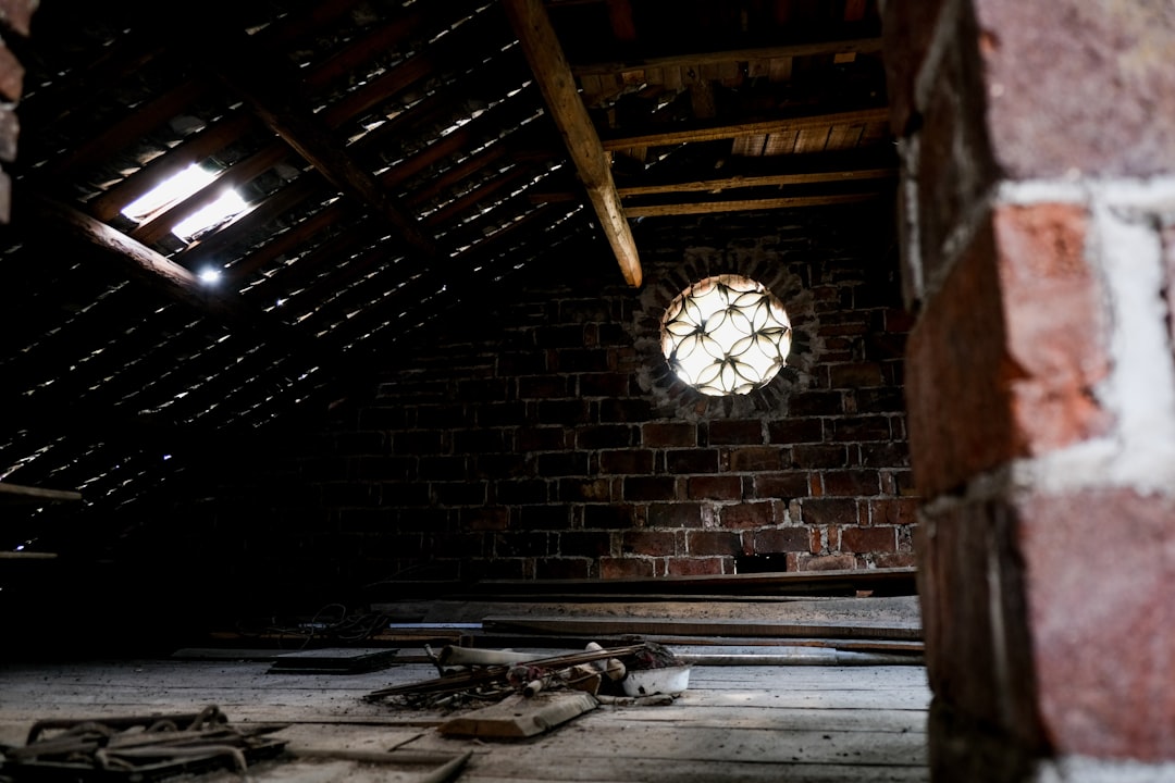 Torn and degraded old insulation in a dark attic, exposing the wooden structural beams behind it and illustrating a primary cause of structural rot.