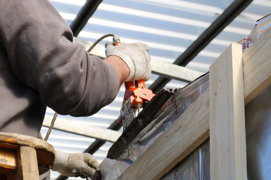 A professional contractor in protective gear is applying expanding spray foam insulation to the underside of a residential roof in an attic, creating an airtight seal.